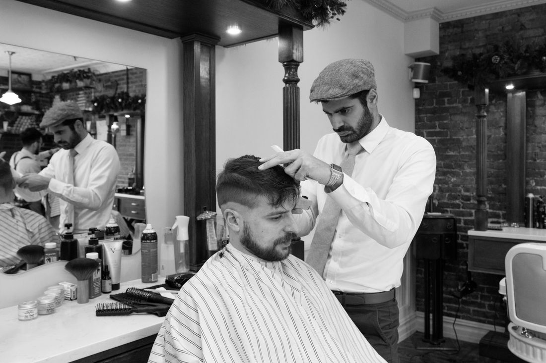 A barber in a white shirt and flat cap cutting a client's hair in a busy shop