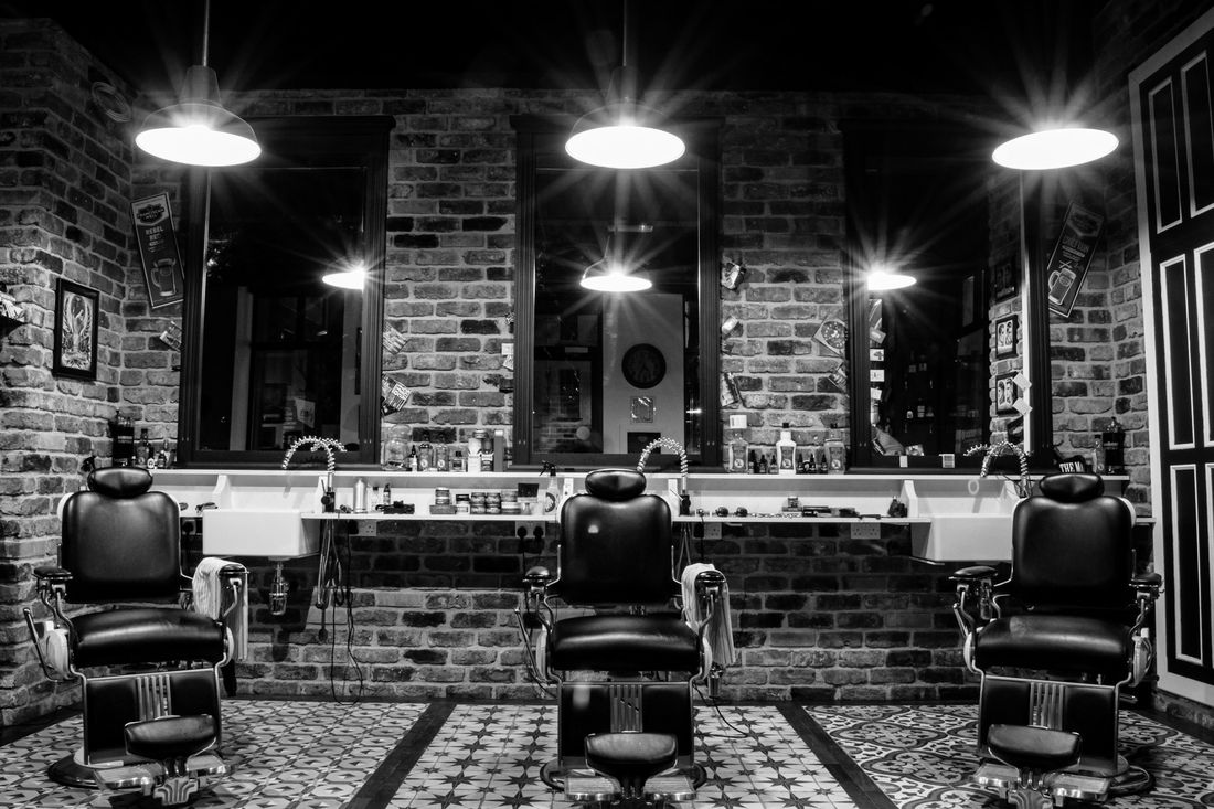 A row of black leather barber chairs facing mirrors in a brick-walled barbershop