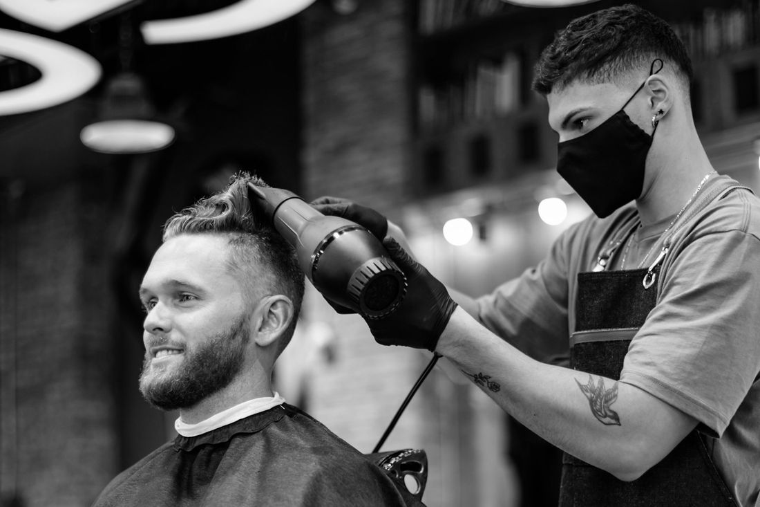 A barber in a face mask styling a client's short hair with a blow-dryer