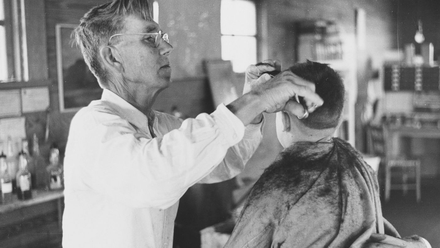 An older barber in a white coat cutting a client's hair in a vintage shop