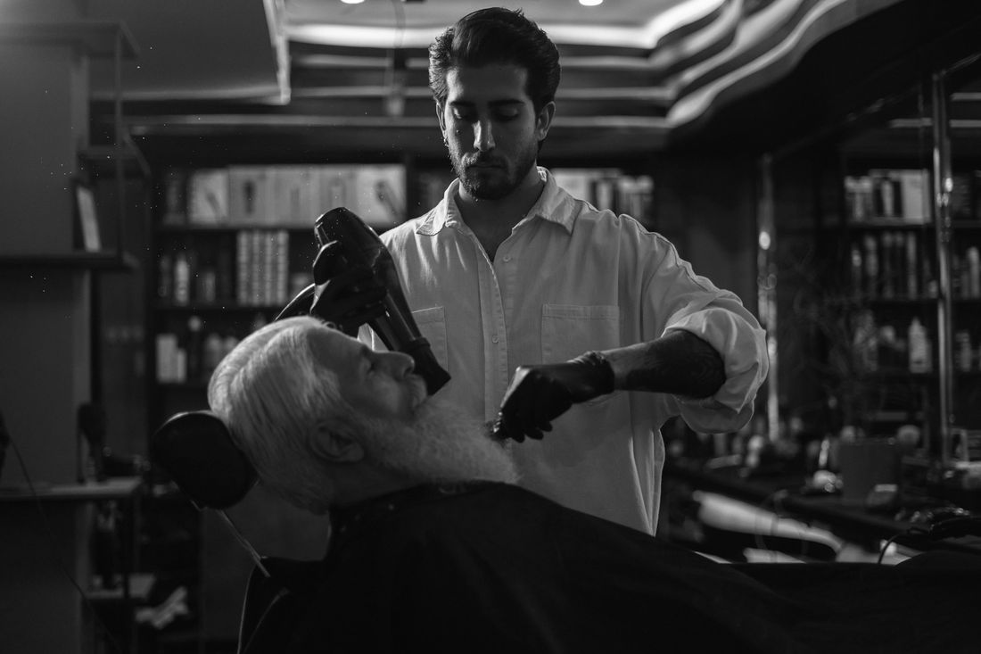 A barber at work on a reclined client in a shop lined with shelves