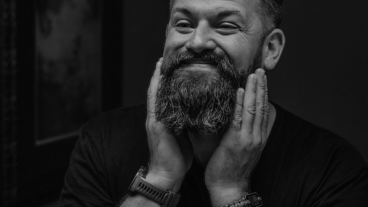 A smiling man with a full, thick beard resting his hands on his cheeks