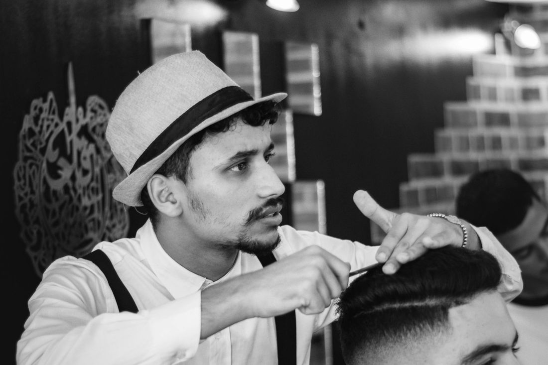 A barber in a brimmed hat cutting a client's hair while talking