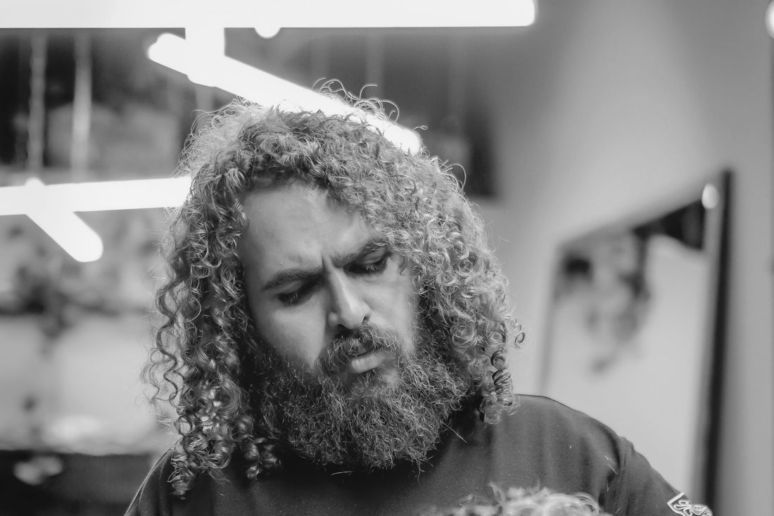 A barber with long curly hair and a full beard at work in his shop
