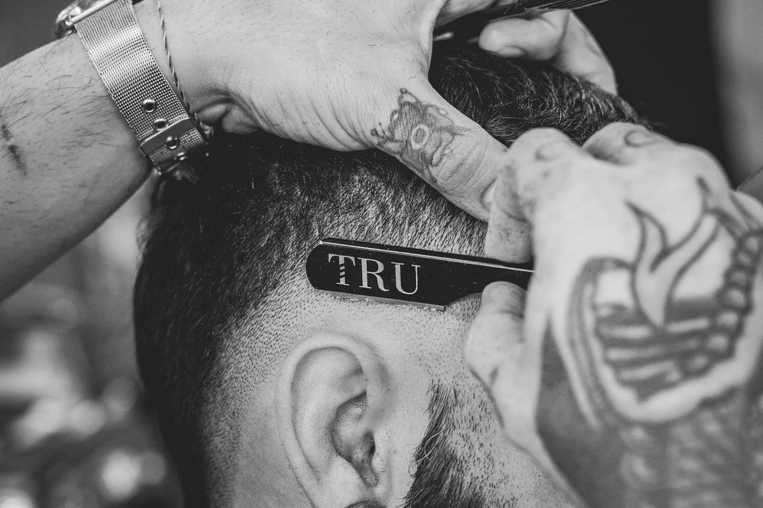 A tattooed barber holding a straight razor against a client's freshly cut temple