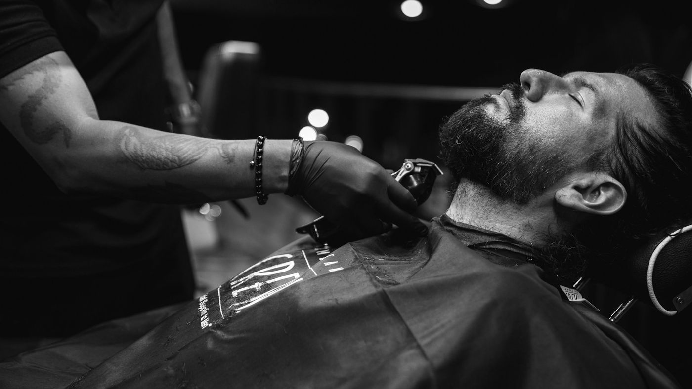 A barber trimming the beard line of a reclining client with a clipper