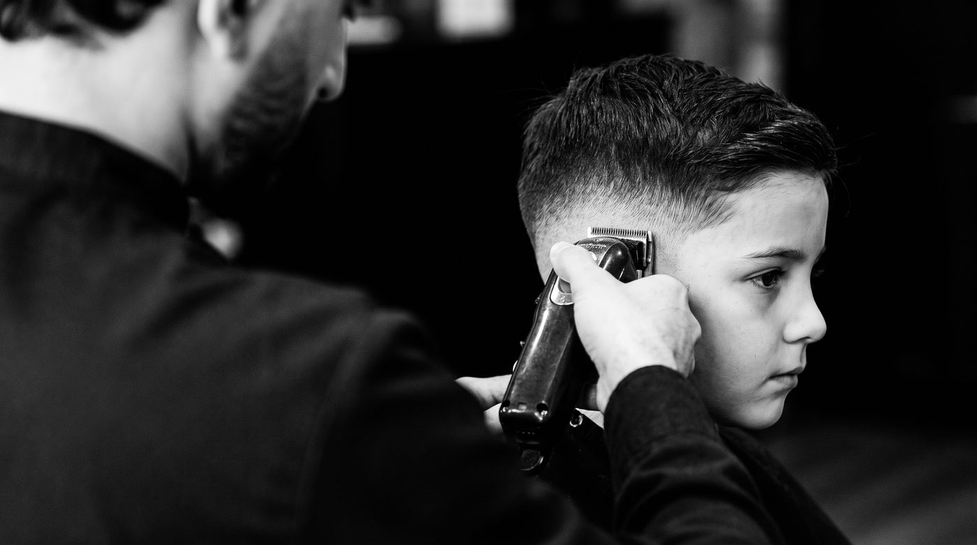 A barber’s hands blending the faded side of a boy’s haircut with clippers, seen from over the shoulder