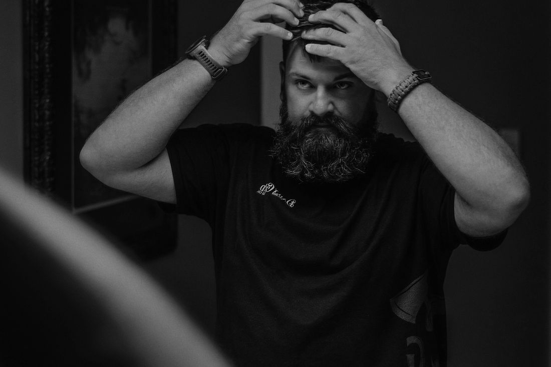 A bearded man looking at himself in a mirror while touching his hair