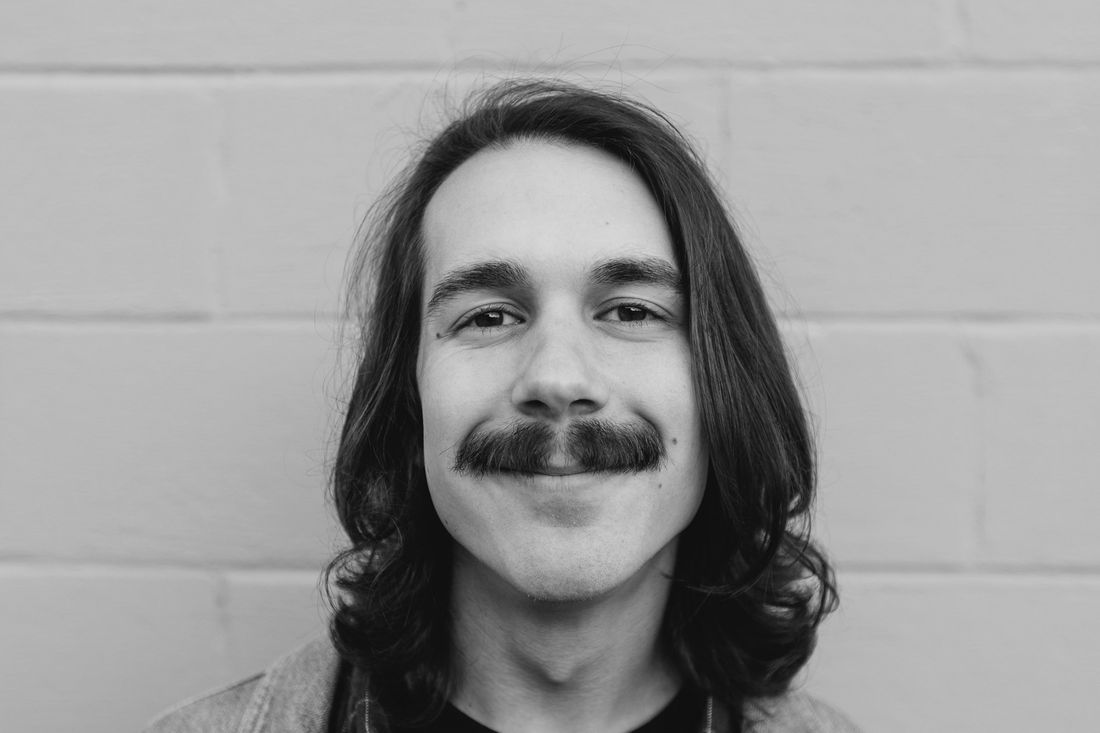 A young man with long hair and a mustache smiling against a white wall