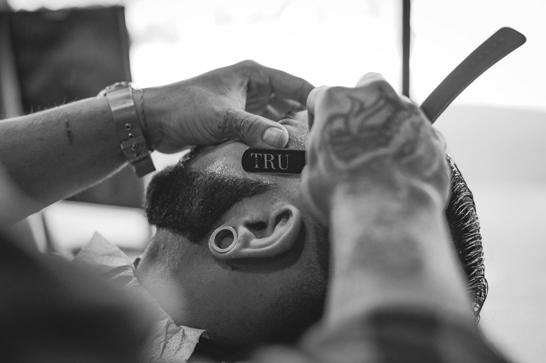 A tattooed barber holding a client's head while working with clippers and a comb