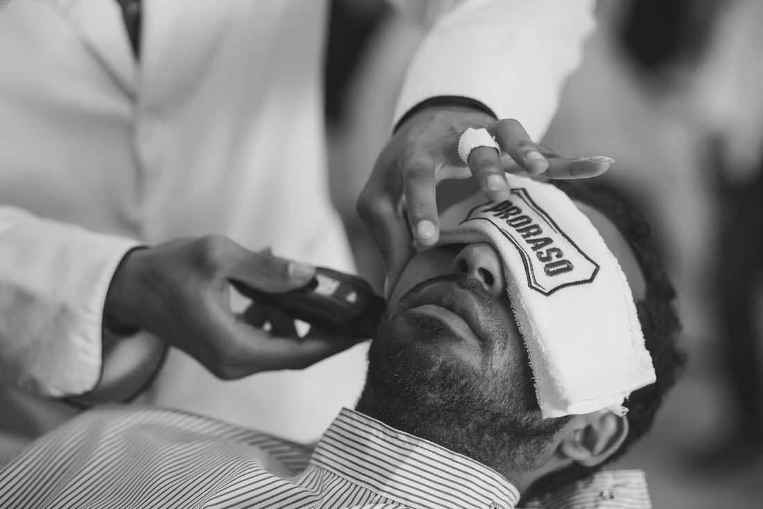 A barber shaving a reclined client with a towel wrapped across his forehead
