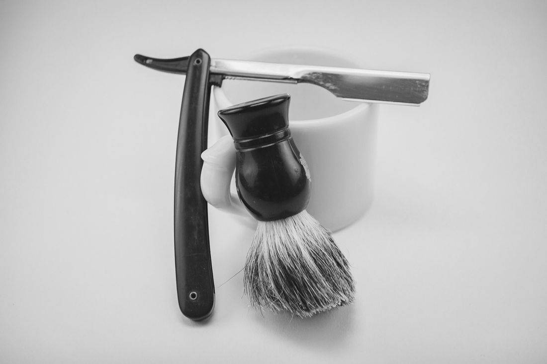 A straight razor, a shaving brush and a ceramic mug of lather on a white surface