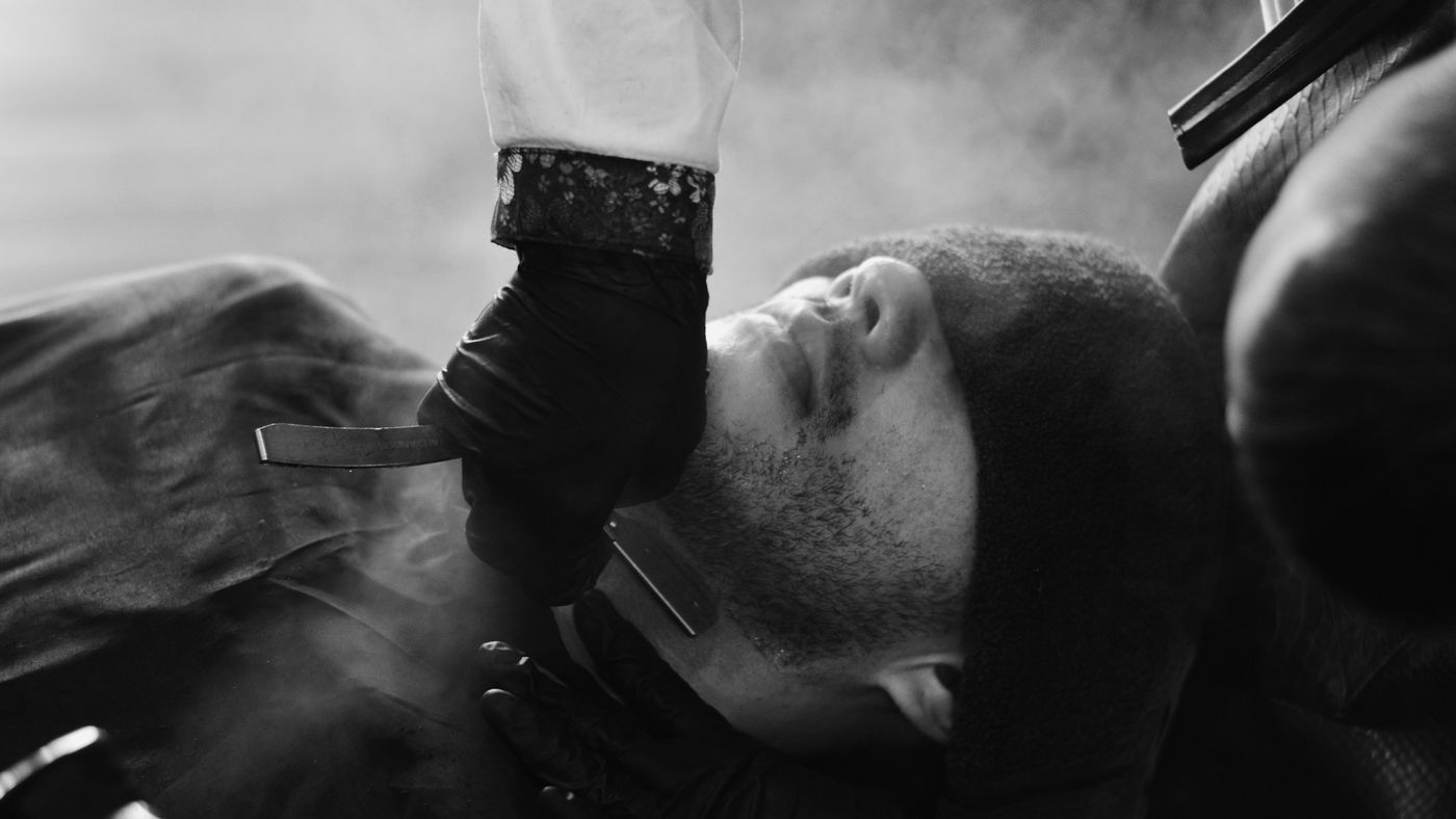 A barber shaving a reclining man's beard with a straight razor