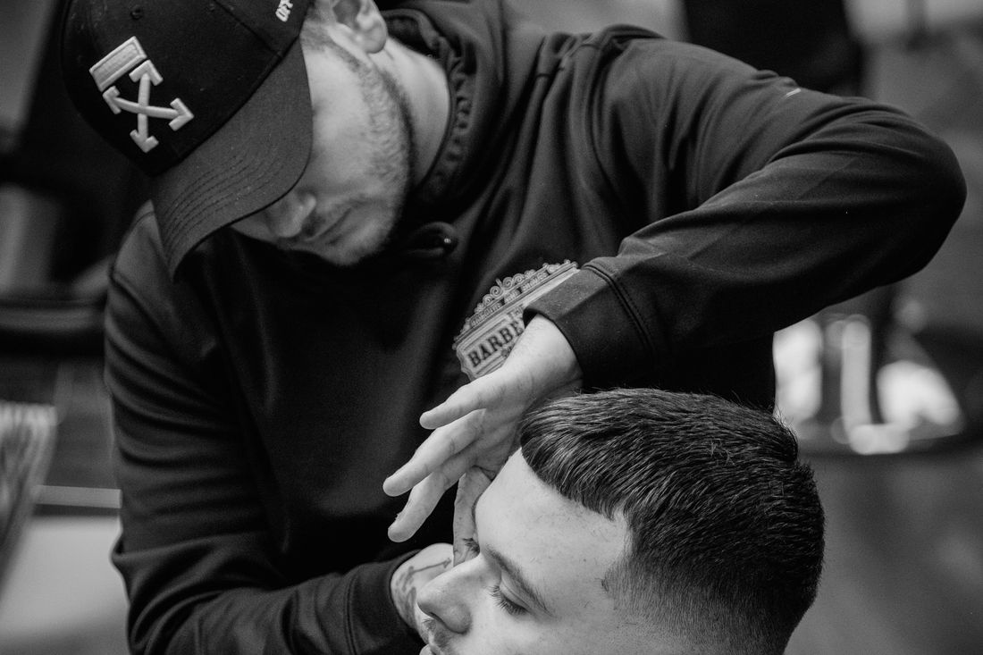 A barber in a cap leaning in to shape a client's hairline