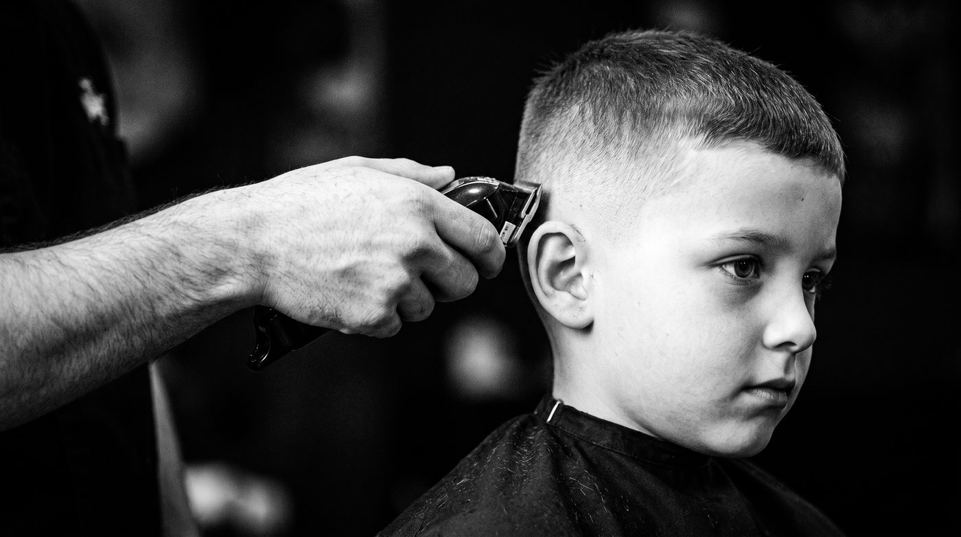 A barber’s cordless clippers held just above a young boy’s ear mid-fade, background softly blurred