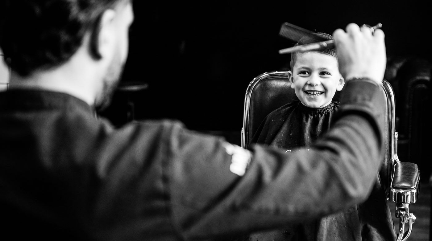 Over the barber’s shoulder: a laughing boy in a barber cape getting a scissor-over-comb trim
