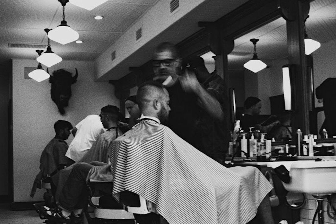 A busy barbershop with clients in several chairs, in black and white