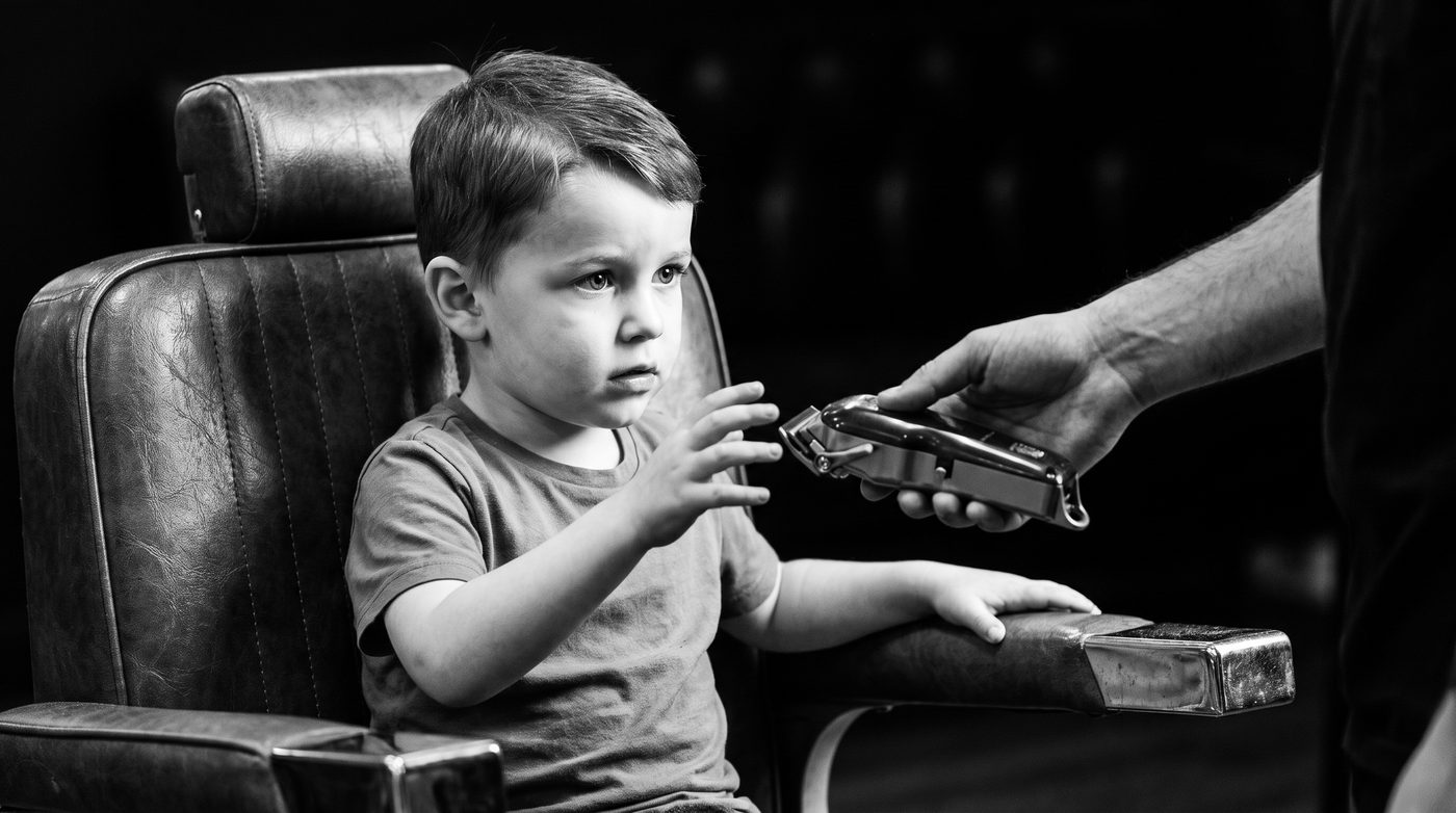 A barber&rsquo;s hand holding out cordless clippers for a young boy in the barber chair to look at and touch
