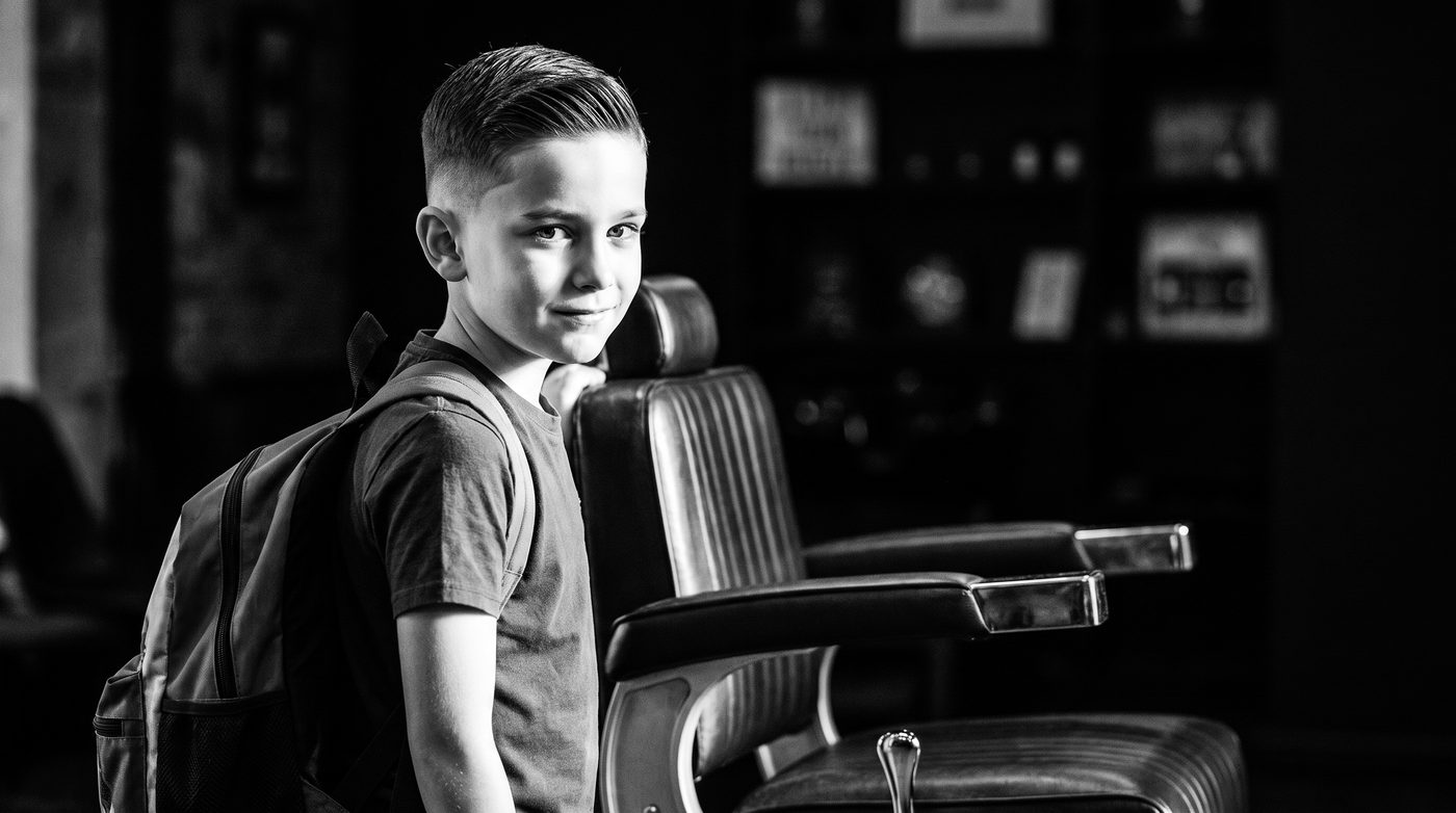 A boy with a fresh sharp haircut and a backpack over one shoulder, lit against the dark of the barbershop