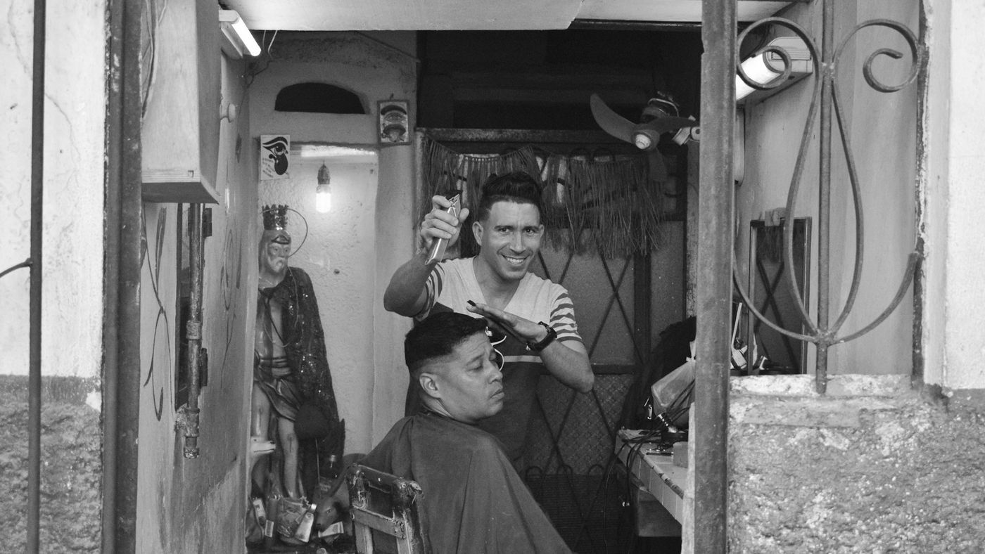 A barber cutting a seated client's hair in a small street-front shop