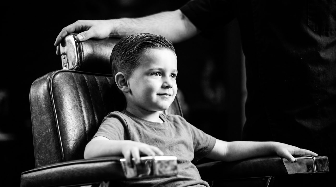 A young boy sitting up straight and proud in a vintage leather barber chair, a barber&rsquo;s hand resting on the chair back